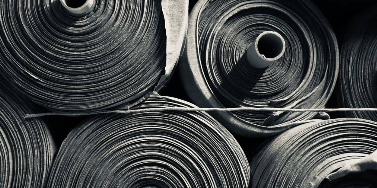 Archivio materico GL MANIFACTORY: prospettiva macro di rotoli di jersey pesante. Studio della densità e della fibra per il Sistema Integrale Herekoretanga. Architettura tessile ispirata al rigore industriale e alla forza della cultura Maori.