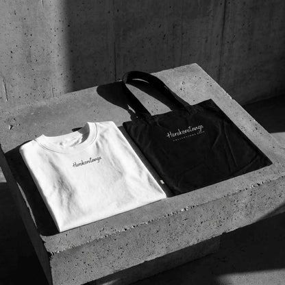 Kit abbigliamento brutalista GL Manifactory composto da t-shirt oversize bianca in heavy jersey di cotone con micro-logo Herekoretanga centrale sul petto e borsa tote bag d'archivio nera in canvas pesante poggiate su blocco di cemento grezzo industriale.
