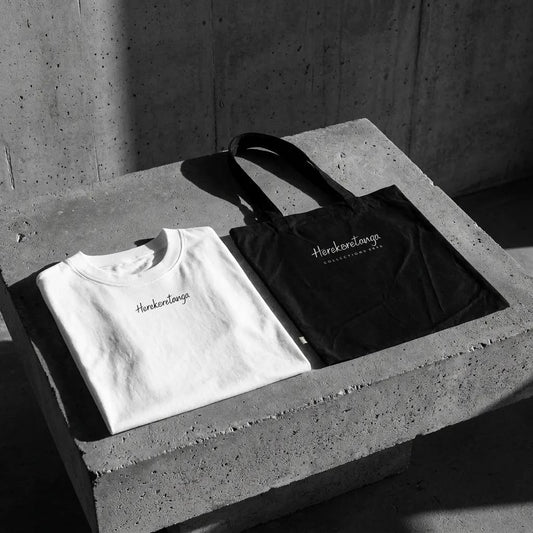 Kit abbigliamento brutalista GL Manifactory composto da t-shirt oversize bianca in heavy jersey di cotone con micro-logo Herekoretanga centrale sul petto e borsa tote bag d'archivio nera in canvas pesante poggiate su blocco di cemento grezzo industriale.
