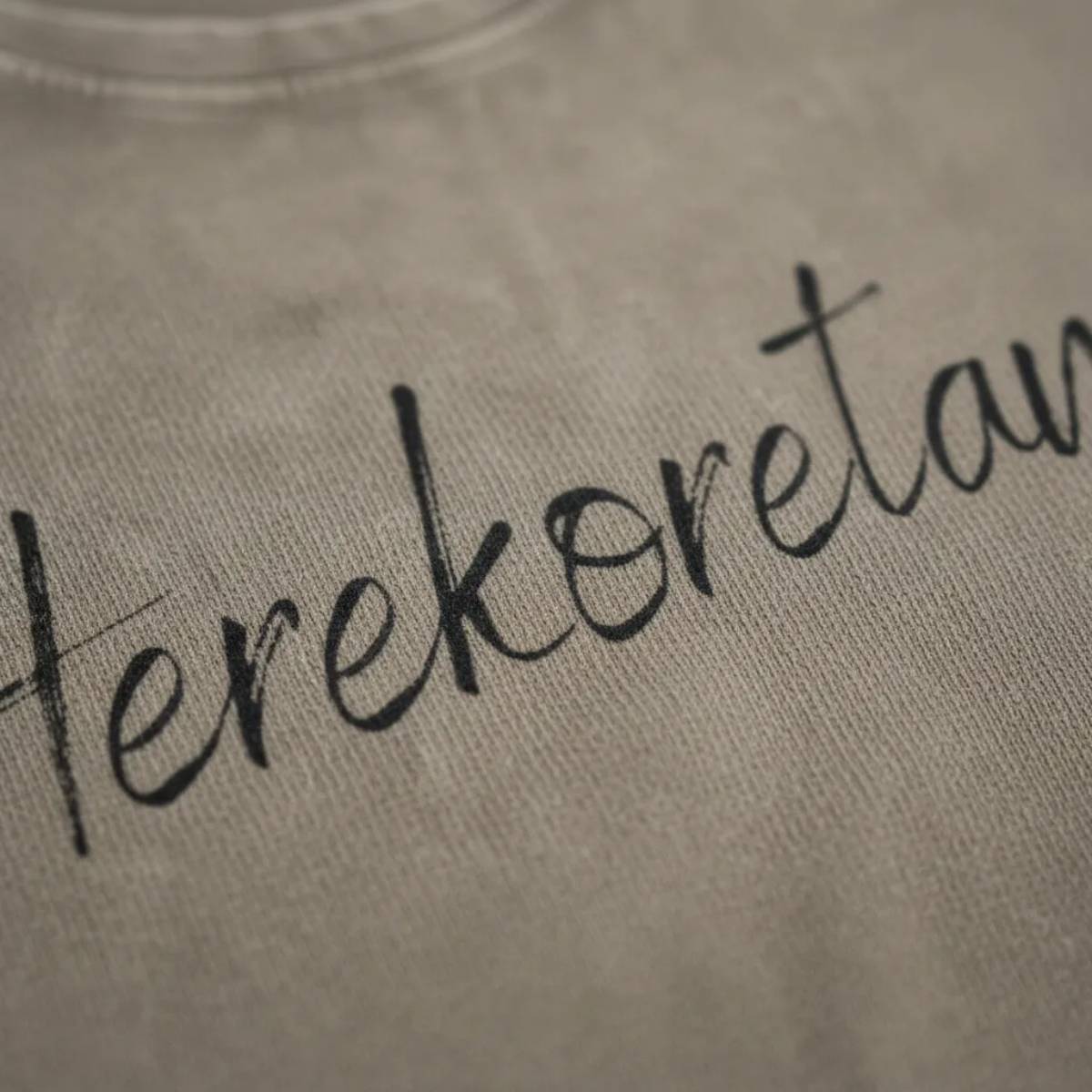 Dettaglio macro della stampa "Herekoretanga" su cotone color kaki scuro. Il close-up esalta la trama del tessuto heavy-duty e la precisione della calligrafia, pilastri del design narrativo GL MANIFACTORY.