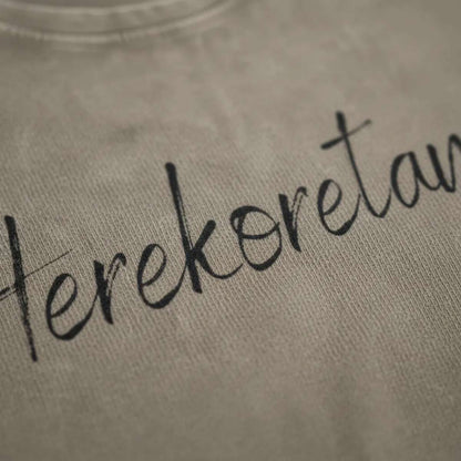 Dettaglio macro della stampa "Herekoretanga" su cotone color kaki scuro. Il close-up esalta la trama del tessuto heavy-duty e la precisione della calligrafia, pilastri del design narrativo GL MANIFACTORY.
