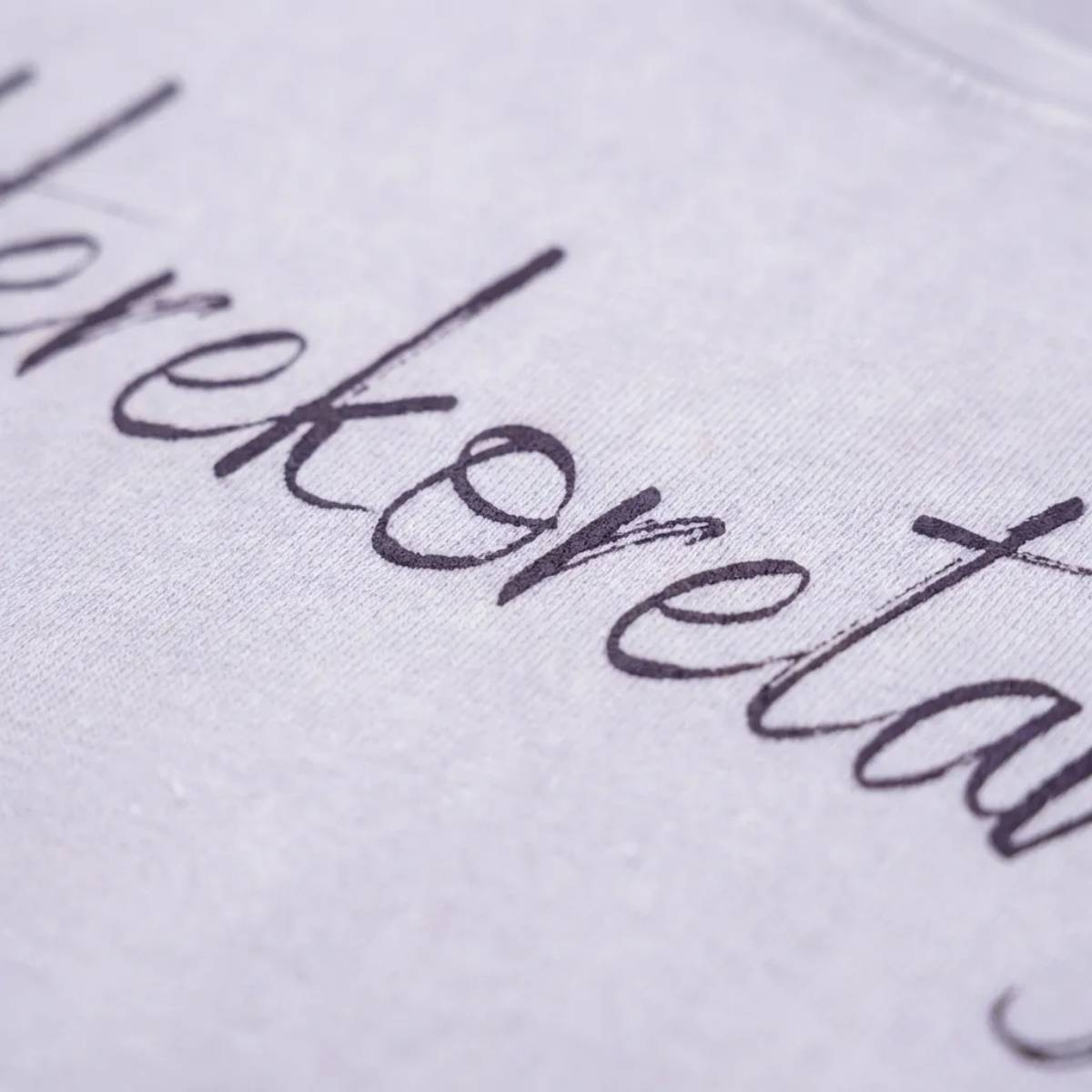 Dettaglio macro della stampa "Herekoretanga" su cotone lilla tenue. Il close-up esalta la trama del tessuto heavy-duty e la precisione della calligrafia, pilastri del design narrativo GL MANIFACTORY.